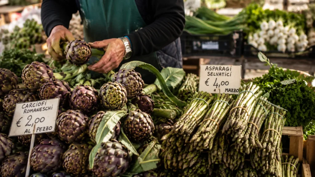 Fresh artichokes and wild asparagus at a Bologna spring market stall, held by a vendor’s hands in natural morning light
